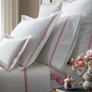 A neatly arranged bedding set with Amara Triple Pearl white sheets and decorative pillows featuring red and pink dotted pearl-like trim, alongside a small bouquet of flowers. The bottom fitted sheet featured belongs to the Charlotte Collection.