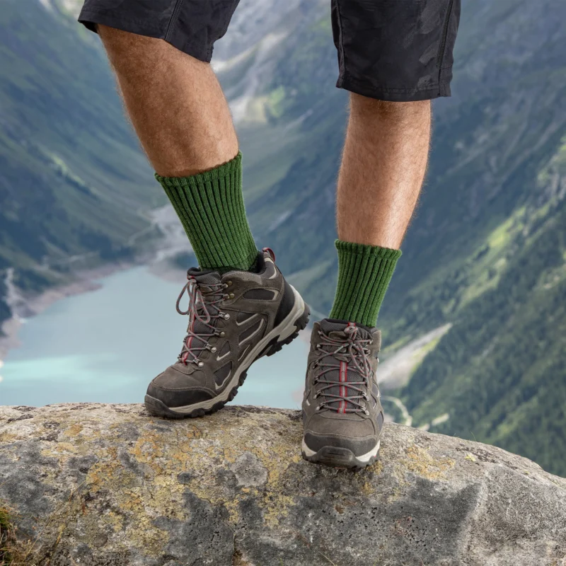 Front view of hiker wearing green ribbed Merino hiking socks with hiking boots, standing on a rock overlooking mountains and lake.