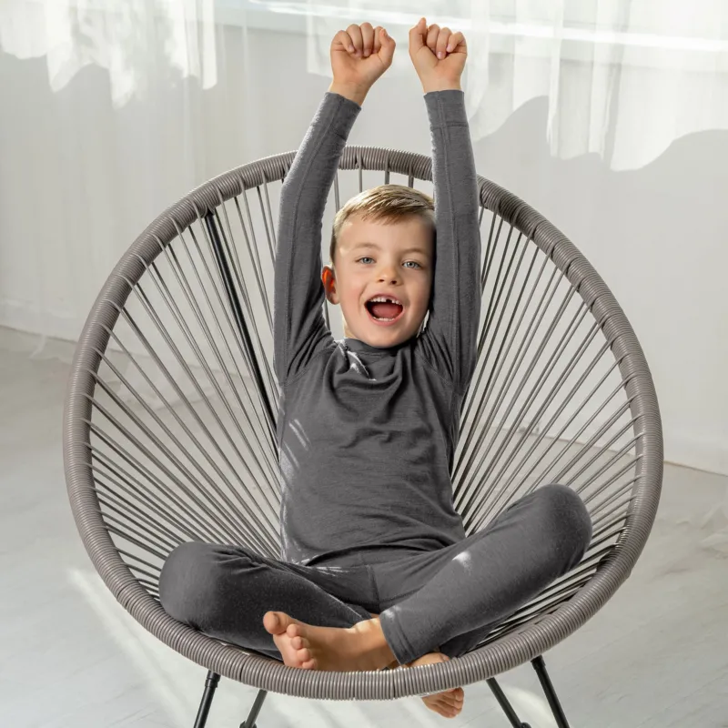 Smiling boy in gray Merino wool base layer set sitting cross-legged on a chair with arms raised.
