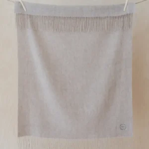 TBCo Lambswool Baby Blanket in Oatmeal Melange with fringed edges hanging on wooden pegs against light wooden background