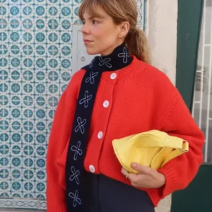 Model wearing a TBCo Merino Wool Skinny Scarf in Navy Flora, adorned with white floral embroidery, layered over a red cardigan, holding a yellow pouch.
