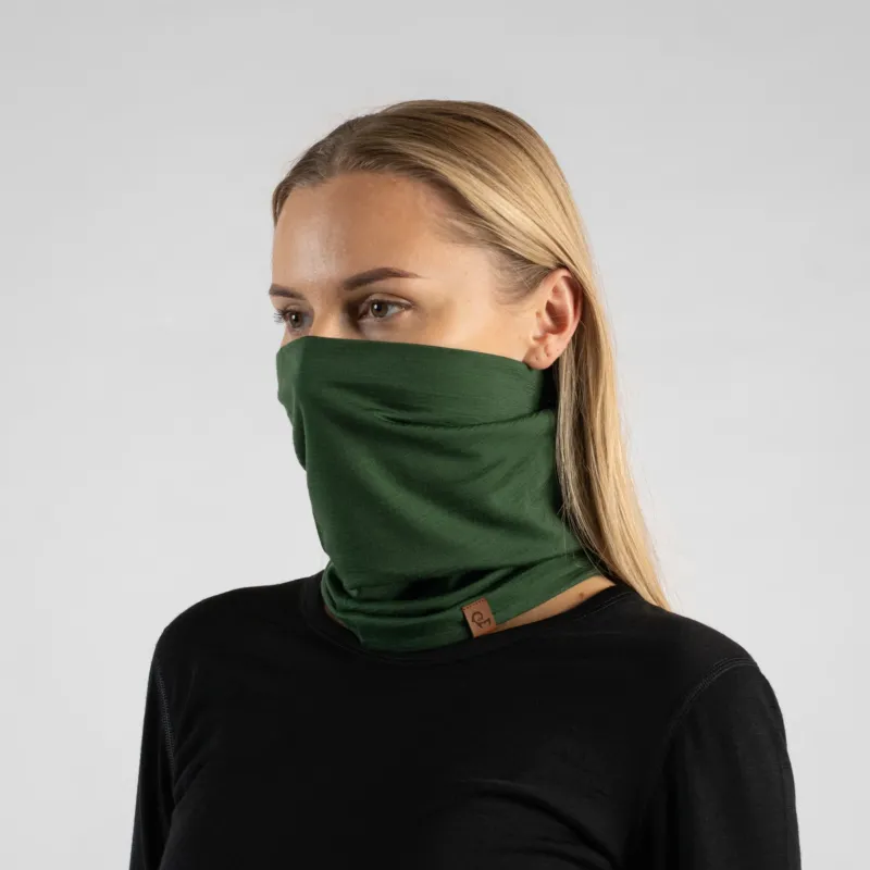 A studio portrait of a blonde woman looking off to the left against a plain light grey background. She is wearing a black long-sleeved shirt and a dark green neck gaiter pulled up to cover her nose and mouth, featuring a small brown logo tag at the bottom.